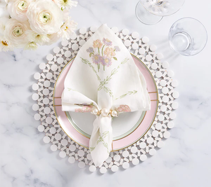The Kim Seybert Trellis Napkin Set, styled with a pearl napkin ring on a pink and white plate and beaded charger, beside glasses and white flowers, makes the perfect spring table accent.