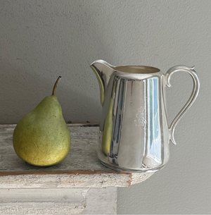 A green pear and the vintage pitcher with Cunard Lines badge from Hôtel Silver are placed side by side on a rustic wooden surface against a plain light gray wall.