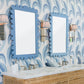 A bathroom features two sinks, two Fleur Home Garden District Coquette Rectangle Mirrors in Denim Wash, wall sconces, and patterned blue wallpaper with feather-like designs inspired by Queen Anne fishtail shingles.