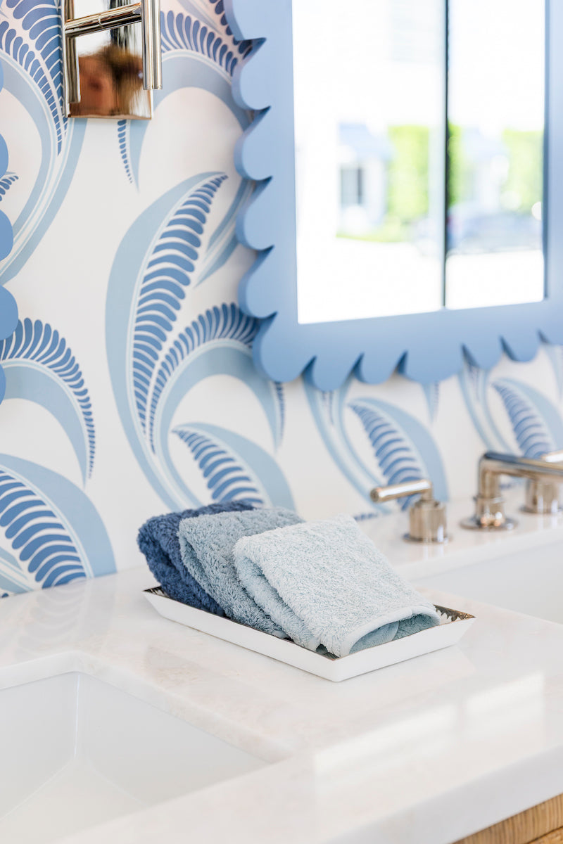 A tray with folded blue washcloths rests on a white countertop near a sink, accented by the Fleur Home Garden District Coquette Rectangle Mirror in Denim Wash and patterned wallpaper inspired by New Orleans charm.