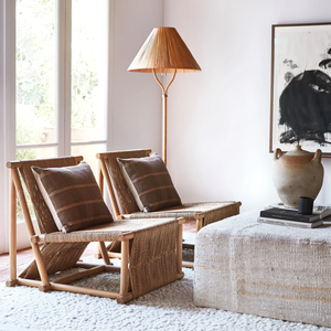 Two woven lounge chairs with brown cushions sit beside the Selamat Delphine Floor Lamp, featuring a handcrafted rattan shade, in a bright living room with a textured ottoman, large vase, brass accents, and abstract art on the wall.