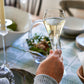 A hand raises a glass from the Simon Pearce Vintner Flute Gift Set above a set dining table, showcasing these elegant flutes—perfect for any celebration.