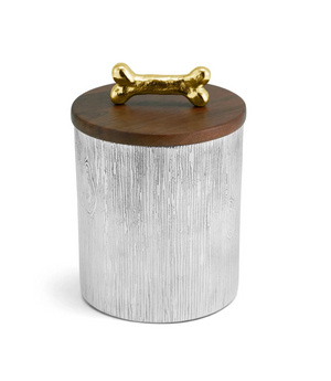 The Dog Bone Canister by Michael Aram features a cylindrical design with a textured silver body, a wooden lid, and a gold bone-shaped handle on top, bringing whimsical charm to any home decor.