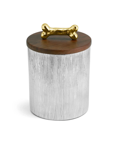 The Dog Bone Canister by Michael Aram features a cylindrical design with a textured silver body, a wooden lid, and a gold bone-shaped handle on top, bringing whimsical charm to any home decor.