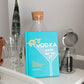 A turquoise cocktail recipe book from Simon & Schuster, titled “Vodka Made Me Do It,” brimming with creative vodka cocktails and infusions, stands on a white cart beside glassware, a plant, and a jar with a cocktail strainer.