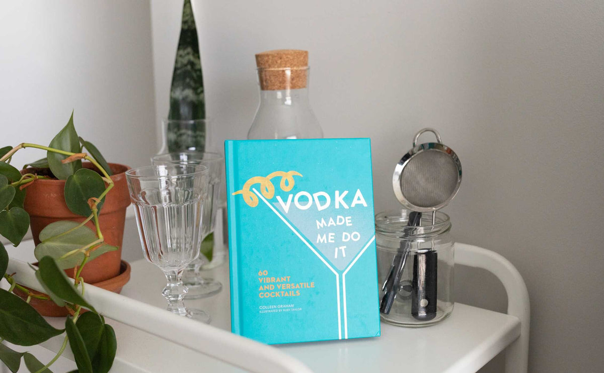 A turquoise cocktail recipe book from Simon & Schuster, titled “Vodka Made Me Do It,” brimming with creative vodka cocktails and infusions, stands on a white cart beside glassware, a plant, and a jar with a cocktail strainer.