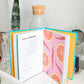 A copy of Vodka Made Me Do It by Simon & Schuster lies open to a “Spicy Spritzer” recipe, its pages adorned with citrus illustrations and surrounded by glassware, a plant, and bottles on a white surface—an inviting scene that inspires you to explore new vodka infusions or craft creative vodka cocktails.