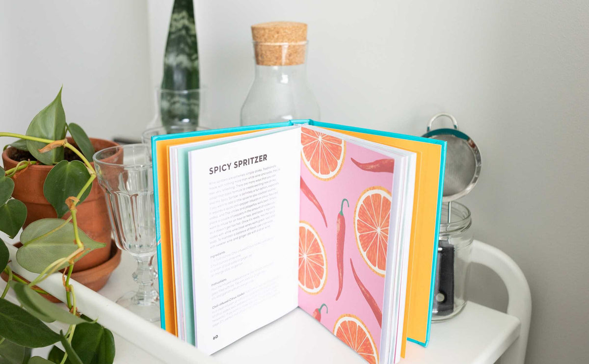 A copy of Vodka Made Me Do It by Simon & Schuster lies open to a “Spicy Spritzer” recipe, its pages adorned with citrus illustrations and surrounded by glassware, a plant, and bottles on a white surface—an inviting scene that inspires you to explore new vodka infusions or craft creative vodka cocktails.