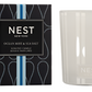 A white Nest Ocean Mist & Sea Salt Candle from the Candle Collection sits by its black and blue box, evoking the refreshing essence of ocean mist with hints of sea salt.