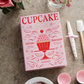 A pink PrintWorks "The Essentials Cupcake Tools" box with cupcake graphics sits on a countertop, accompanied by a spatula, rolled napkin with ribbon, flowers, and other baking essentials.