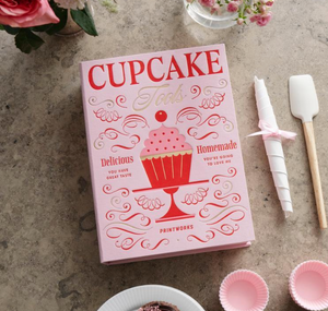 A pink PrintWorks "The Essentials Cupcake Tools" box with cupcake graphics sits on a countertop, accompanied by a spatula, rolled napkin with ribbon, flowers, and other baking essentials.