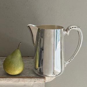 A green pear rests beside the Hôtel Silver Vintage Pitcher with Perforated Handles on a worn wooden surface, set against a light gray wall.