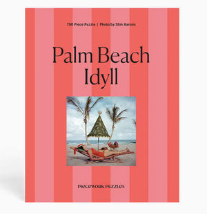 A 750-piece puzzle from the Piecework Slim Aarons Puzzle Collection features the “Palm Beach Idyll” photo of people relaxing under a canopy on a beach with palm trees, set against a pink and red striped background, and is printed on high-quality art paper for vibrant colors and durability.