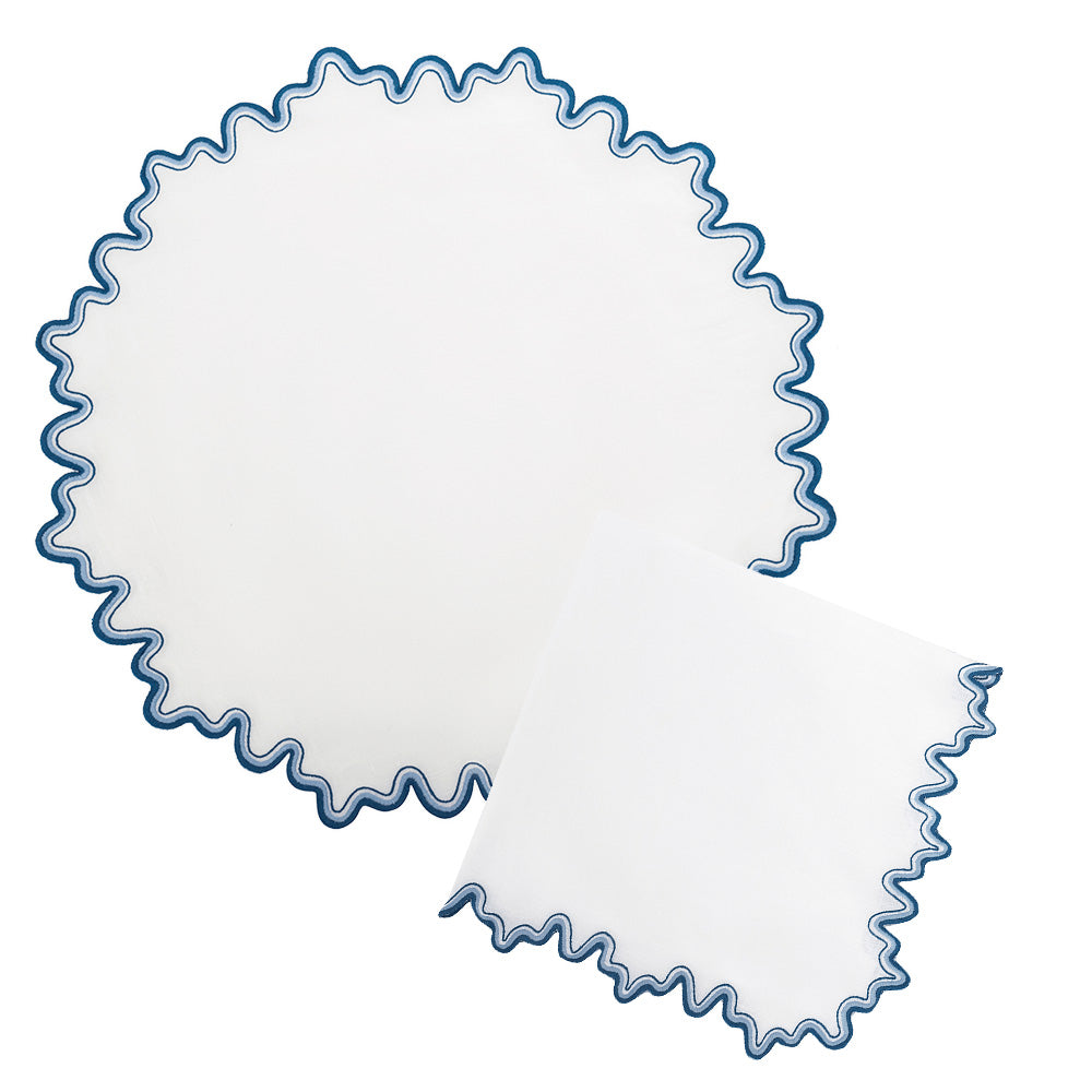 Matouk's Wave Tabletop Collection, Aquarius, features white round and square linen napkins with blue ruffled edges, OEKO-TEX Standard 100 certified for safety. The round napkin is laid flat and the square napkin is folded, adding embroidered charm to your table.