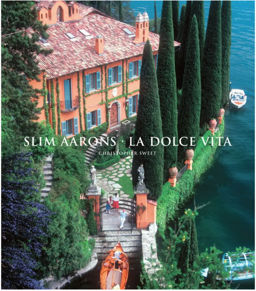 A sun-drenched villa with blue shutters overlooks a lake in the Slim Aarons: La Dolce Vida photography, showing people in a small boat and near a gate.