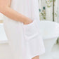 A person wearing a Weezie Towels White Towel Wrap with a pocket stands hands-free by a white bathtub in a bathroom with floral curtains.