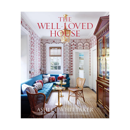 Book cover of The Well-Loved House from Penguin Random House, showcasing a vibrant, traditional living room with patterned wallpaper and a blue sofa—a perfect representation of the neo-traditionalist interior design style that defines Ashley Whittaker’s work.