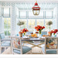 A luminous dining room—featuring a rectangular table arranged for six, floral-upholstered chairs, blue accents, vivid red flowers, a striking red lantern, and expansive windows streaming in daylight—showcases the neo-traditionalist interior design that defines Ashley Whittaker’s distinctive approach in The Well-Loved House by Penguin Random House.