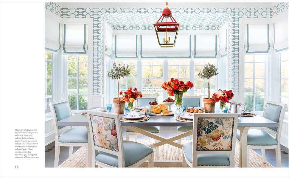 A luminous dining room—featuring a rectangular table arranged for six, floral-upholstered chairs, blue accents, vivid red flowers, a striking red lantern, and expansive windows streaming in daylight—showcases the neo-traditionalist interior design that defines Ashley Whittaker’s distinctive approach in The Well-Loved House by Penguin Random House.