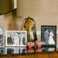A table displays Addison Ross’s Enamel Locket Frame with a 5x7 inch black-and-white wedding photo, two pink decor pieces, and a brown glass lamp in the background for a timeless, elegant touch.