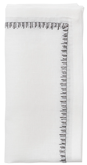 The Filament Napkin Set by Kim Seybert features crisp white linen napkins, elegantly folded and accented with decorative black embroidered stitching along two edges.