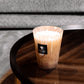 A Baobab Collection Pearls White Candle scented candle in a glass container placed on a wooden coffee table, with a gray couch in the background.