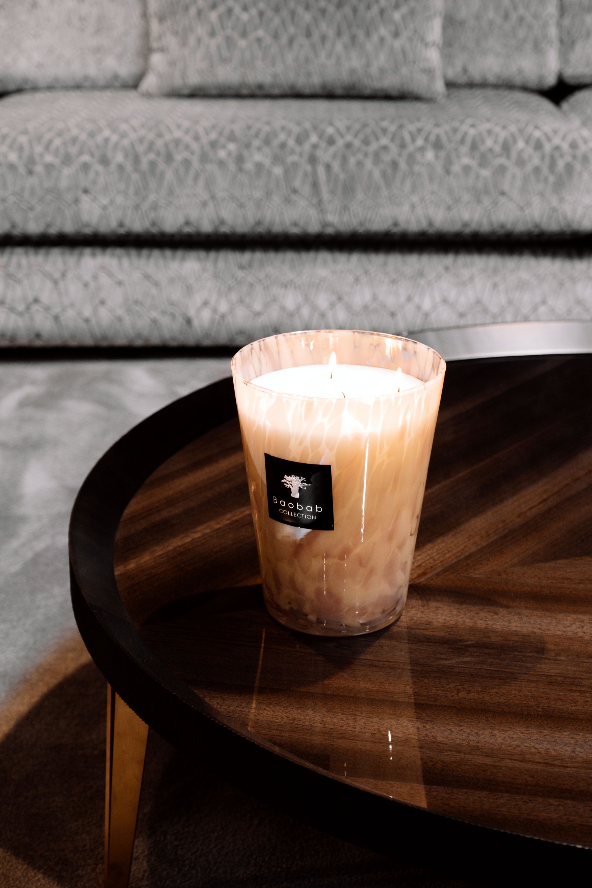 A Baobab Collection Pearls White Candle scented candle in a glass container placed on a wooden coffee table, with a gray couch in the background.