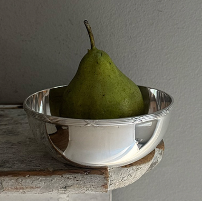 A green pear sits upright in a Hôtel Silver Vintage Bowl with Ribbon & Reed Top Rim, placed on the corner of a rustic wooden surface against a gray wall.