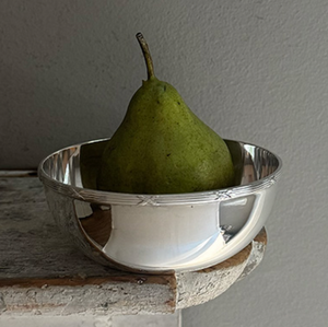 A green pear sits upright in a Hôtel Silver Vintage Bowl with Ribbon & Reed Top Rim, placed on the corner of a rustic wooden surface against a gray wall.