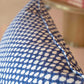 Close-up of the Royal Blue Wicker Outdoor Pillow by Associated Design features a blue and white pattern with visible edge stitching; a blurred table with books appears in the background.