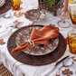 Elegant autumn table setting featuring Kim Seybert's Wood Matrix Placemat, brown floral plates, an orange napkin with a ring, amber glasses, silver cutlery, and a white tablecloth with leaf embroidery.