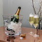 A Simon Pearce Woodbury Chiller holds a bottle of champagne next to two filled flutes and opened corks on a copper-colored table.