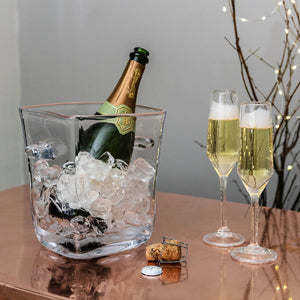 A Simon Pearce Woodbury Chiller holds a bottle of champagne next to two filled flutes and opened corks on a copper-colored table.