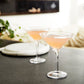Two pink cocktails in contemporary glasses from the Simon Pearce Woodbury Martini Glass Gift Set sit on a gray countertop, with greenery and neutral decor blurred in the background—showcasing this elegant barware.