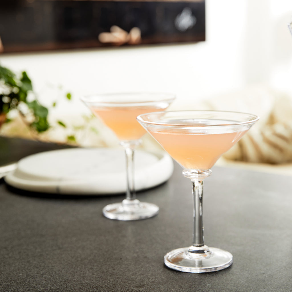 Two pink cocktails in contemporary glasses from the Simon Pearce Woodbury Martini Glass Gift Set sit on a gray countertop, with greenery and neutral decor blurred in the background—showcasing this elegant barware.
