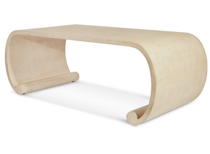 The Emilia Coffee Table by Bunny Williams features a beige, modern design with a smooth surface, curved sides forming partial loops, and a stylish painted linen finish inspired by Ming dynasty furniture.