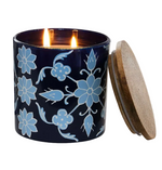 Lit two-wick Goa Candle & Incense Collection ylang-ylang candle in a dark blue jar with a light blue floral pattern, sitting next to its wooden lid.