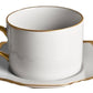 White porcelain teacup with a gold handle and rim, set on a matching saucer adorned with a scalloped gold-edged border, from the Anna’s Palette Antique White with Gold Dinnerware Collection by Anna Weatherley.
