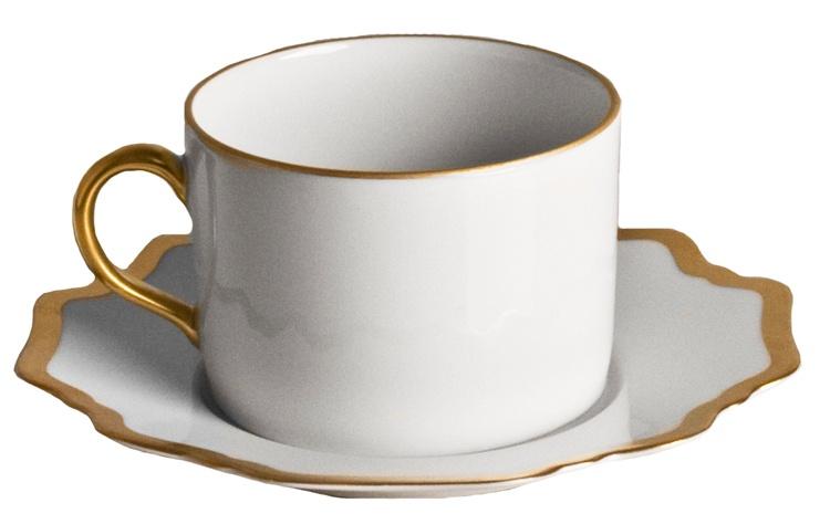 White porcelain teacup with a gold handle and rim, set on a matching saucer adorned with a scalloped gold-edged border, from the Anna’s Palette Antique White with Gold Dinnerware Collection by Anna Weatherley.