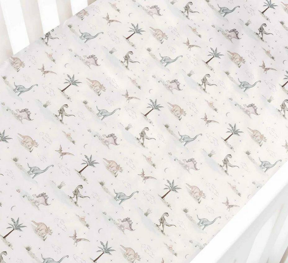 The Gooselings Dino Dreams Bedding Collection features a white crib sheet adorned with illustrated dinosaurs, palm trees, and rocks in soothing neutral tones. Expertly fitted over a crib mattress, this sheet is crafted from breathable fabric for optimal baby comfort and is OEKO-TEX certified for added peace of mind.