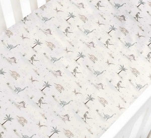 The Gooselings Dino Dreams Bedding Collection features a white crib sheet adorned with illustrated dinosaurs, palm trees, and rocks in soothing neutral tones. Expertly fitted over a crib mattress, this sheet is crafted from breathable fabric for optimal baby comfort and is OEKO-TEX certified for added peace of mind.
