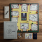 A Clue Vintage Bookshelf Edition board game set by WS Game Company on a wooden table, featuring a game board, clue cards, hall, lounge, ballroom, other rooms, tokens, score sheets, a linen-wrapped booklet from the Hasbro editions collection, and dice.