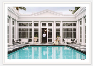 A person in formal attire stands at the edge of a pool with tall white columns and large windows in the background, reminiscent of Palm Beach elegance, holding a Nick Mele Fine Art Bunny Ears from Nick Mele Photography, while another sits on a bench by the poolside.
