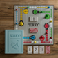 A Sorry! Vintage Bookshelf Edition board game set up on a wooden surface, featuring pawns, cards, and a game board with pathways and marked spaces. A blue SORRY! Vintage Bookshelf Edition game box from WS Game Company is placed next to the vintage game board, adding charm and nostalgia.