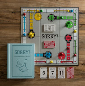 A Sorry! Vintage Bookshelf Edition board game set up on a wooden surface, featuring pawns, cards, and a game board with pathways and marked spaces. A blue SORRY! Vintage Bookshelf Edition game box from WS Game Company is placed next to the vintage game board, adding charm and nostalgia.