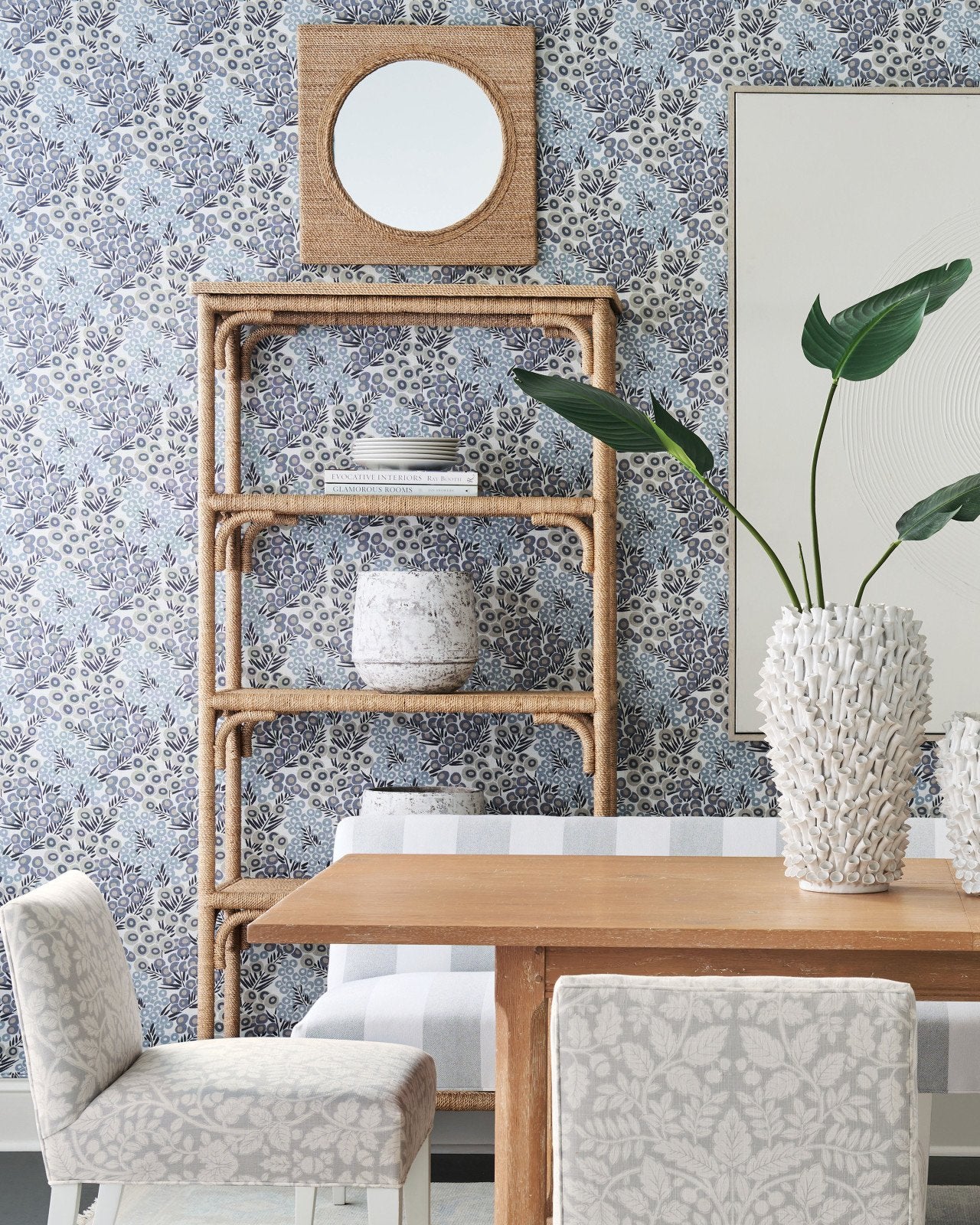 Dining area with a wooden table, upholstered chairs, and the Currey and Company Olisa Etagere displaying decorative vases, a leafy plant, and set against floral-patterned wallpaper. A round mirror hangs above the shelves.
