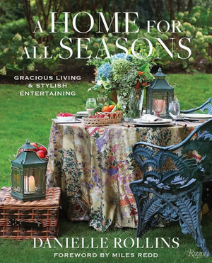 An elegantly styled outdoor dining table—adorned with a floral tablecloth, coordinated plates, glassware, and lanterns—rests on a lush lawn surrounded by garden chairs. The inviting scene beautifully reflects the art of stylish entertaining, while the cover title “A Home For All Seasons” from Penguin Random House overlays the image, evoking Danielle Rollins’s signature approach to refined home decor.