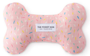 A pink, bone-shaped toy from the Dog Bone Squeaky Toy Collection by Foggy Dog, featuring multicolored sprinkles and a center label that reads "The Foggy Dog San Francisco," handcrafted for quality.