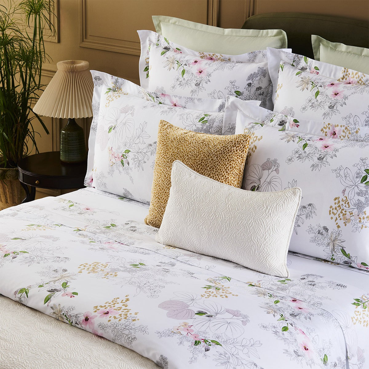 A neatly made bed features the Yves Delorme Orchidee Bedding Collection in organic cotton sateen, paired with multiple pillows and a beige accent pillow, set beside a lamp, green plant, and paneled wall.