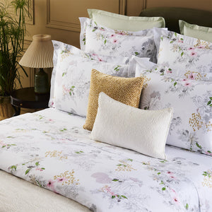 A neatly made bed features the Yves Delorme Orchidee Bedding Collection in organic cotton sateen, paired with multiple pillows and a beige accent pillow, set beside a lamp, green plant, and paneled wall.